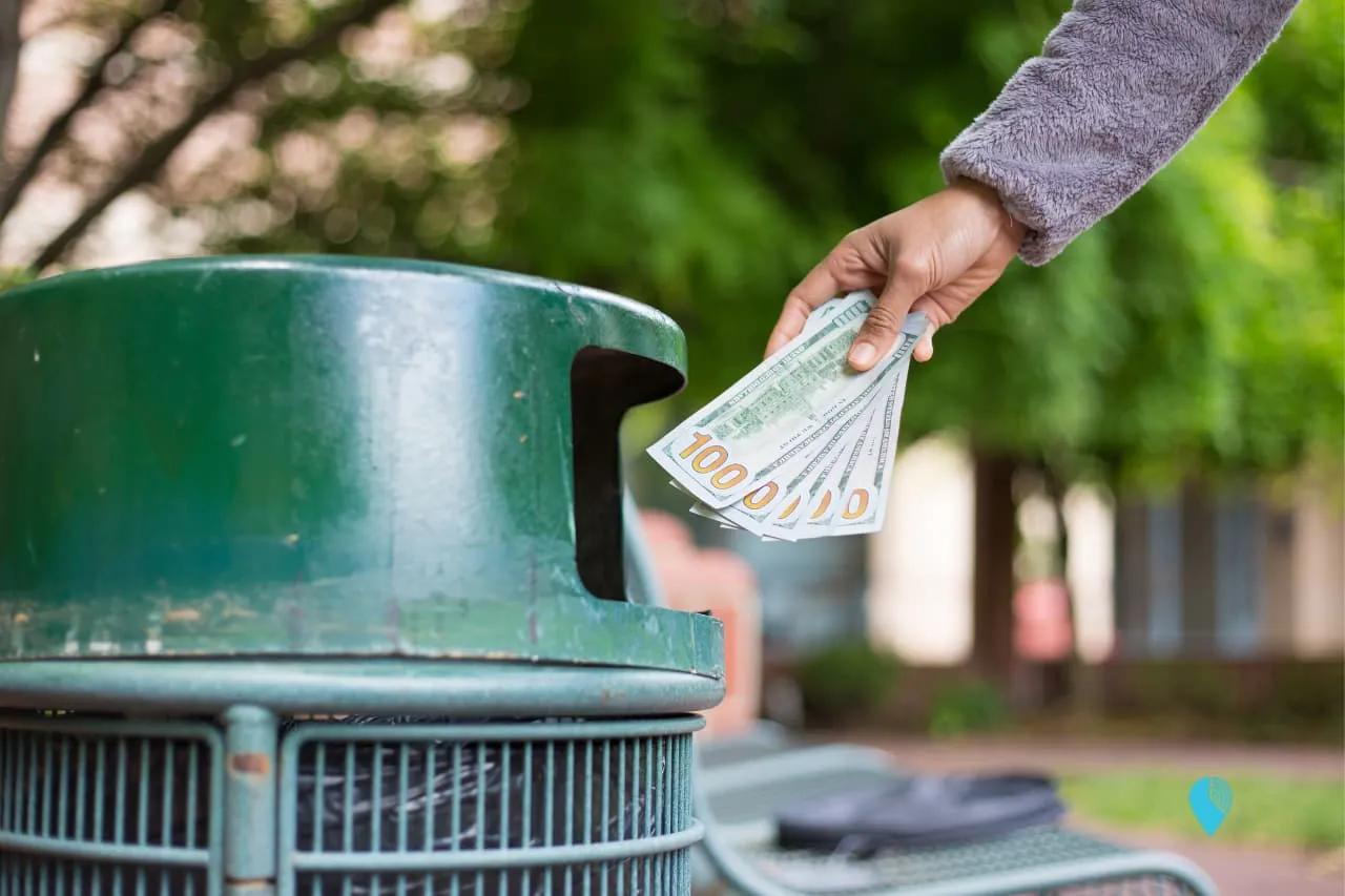 a man throwing several $100 bills into the trash can representing money wasted manually converting hours minutes to decimal time
